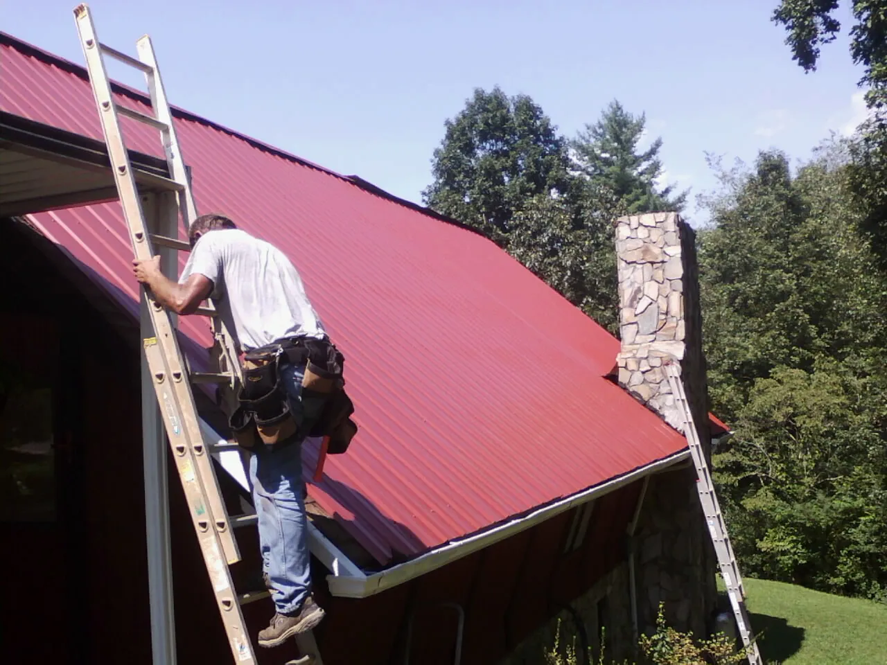 Roofer on a ladder beside a red metal roof and stone chimney — Soffit & Fascia Repair by Roofing Brothers in Shattuck